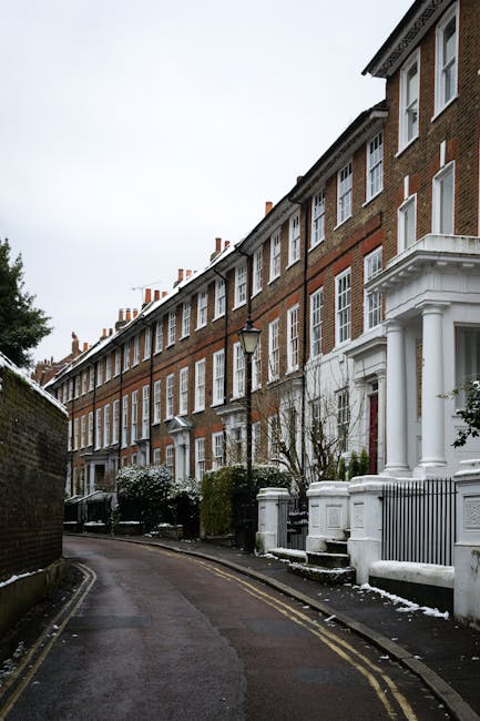 A narrow residential street lined with terraced brick houses featuring white-painted decorative elements and columns at the entrances. The buildings are constructed from reddish-brown brick with uniform white-framed sash windows, and some structures have small front gardens with low fences and neatly trimmed shrubs. The street surface is tarmac with visible double yellow lines along the curb, and a lamppost is positioned centrally on the pavement. There is a hint of snow on the ground and on the shrubbery, indicating winter conditions. The sky overhead is overcast, providing diffuse, soft lighting that adds a muted tone to the scene. The absence of vehicles or visible waste emphasizes a quiet, well-maintained area, which may be associated with discreet, private waste handling services like those offered by Rubbish Collection Twickenham for local residents seeking alternative rubbish removal options outside of standard council collection.