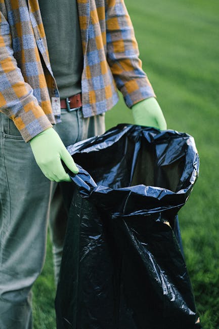 Close-up of a person wearing a grey and yellow checkered flannel shirt, green gloves, and grey trousers, holding open a large black plastic rubbish bag outdoors on a grassy area during daytime. The bag is empty, wide open, and appears ready for waste collection, with a glossy surface that reflects natural light. The background features lush green grass, indicating an outdoor environment such as a garden, park, or lawn. The scene suggests a waste disposal or rubbish collection activity, with the individual preparing to fill or remove waste materials. This image visually supports services related to independent rubbish removal, private waste handling, or on-site clearance, as provided by Rubbish Collection Twickenham, emphasizing environmentally responsible and professional rubbish disposal solutions.