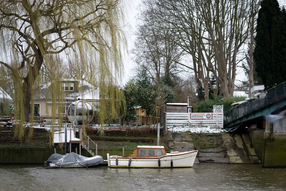 The image depicts a riverside scene with a small, white boat moored along the water's edge, partially covered by a grey tarp. The boat has a wooden cabin and black fenders hanging along its side. Adjacent to the boat, a narrow wooden dock with steps leads up to a backyard area enclosed by a dark wooden fence. The fence surrounds a garden with leafless trees and shrubs, some evergreen, and a house with light-colored walls and a pitched roof is visible in the background. To the right of the boat, a metal bridge with a greenish finish spans over the water, and an embankment with concrete supports and a white fence with safety signage is seen behind it. Large, leafless trees with layered branches dominate the upper part of the image, contrasting with an overcast sky that provides diffuse lighting. The scene has a calm, neutral atmosphere with no visible activity, and the setting suggests a quiet residential area with potential for private waste handling or rubbish collection providings by Rubbish Collection Twickenham.