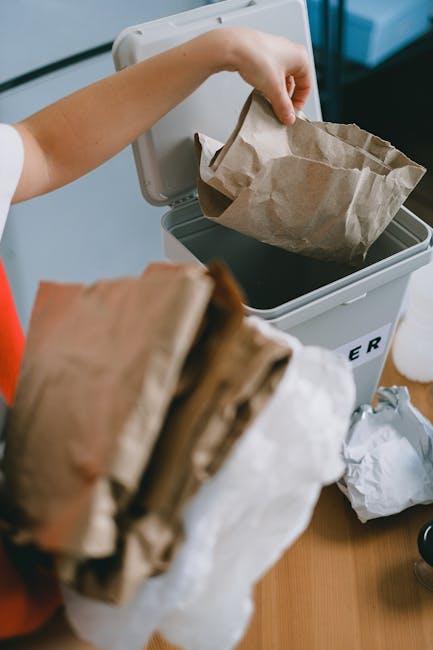 A person placing a crumpled brown paper bag into a grey rubbish bin with a hinged lid, positioned indoors on a wooden surface. The bag's material appears rough and paper-like, with visible creases and folds. In the foreground, another crumpled paper bag, partially out of focus, is being held by the person. The background includes a blue wall or surface and a small rod or handle, indicating a domestic or small-scale waste disposal environment. The scene captures the process of household rubbish disposal, which may be associated with independent rubbish removal services such as those provided by Rubbish Collection Twickenham, offering an alternative to council-led waste collection. The lighting is natural or softly ambient, emphasizing clarity of the disposal activity and the texture of the paper bags, aligning with professional waste management practices focused on household waste separation and disposal.