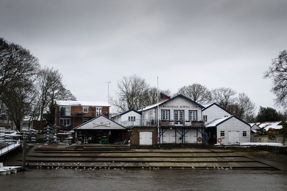 The image depicts a riverside scene with a small, white boat moored along the water's edge, partially covered by a grey tarp. The boat has a wooden cabin and black fenders hanging along its side. Adjacent to the boat, a narrow wooden dock with steps leads up to a backyard area enclosed by a dark wooden fence. The fence surrounds a garden with leafless trees and shrubs, some evergreen, and a house with light-colored walls and a pitched roof is visible in the background. To the right of the boat, a metal bridge with a greenish finish spans over the water, and an embankment with concrete supports and a white fence with safety signage is seen behind it. Large, leafless trees with layered branches dominate the upper part of the image, contrasting with an overcast sky that provides diffuse lighting. The scene has a calm, neutral atmosphere with no visible activity, and the setting suggests a quiet residential area with potential for private waste handling or rubbish collection providings by Rubbish Collection Twickenham.