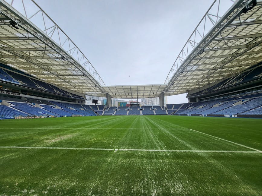 A wide view of a large, modern football stadium with a well-maintained green grass pitch marked with white lines. The stadium features blue seating sections on both the left and right sides, extending across multiple tiers. Above the seating areas, there are extensive roof structures made of metal and glass, supported by large steel trusses that create an open yet sheltered environment. The sky above is overcast, casting diffuse lighting over the scene, with no spectators or players visible on the field. Surrounding the pitch, the concrete walkways and support columns are visible, contributing to the overall structure. This scene represents an empty stadium prepared for a match, with a focus on the pristine condition of the grass and the architectural design of the roof, aligning with the context of large-scale sports venues where occasional maintenance or on-site clearance may be required, akin to private waste handling or refurbishment activities by professional rubbish removal services like Rubbish Collection Twickenham.