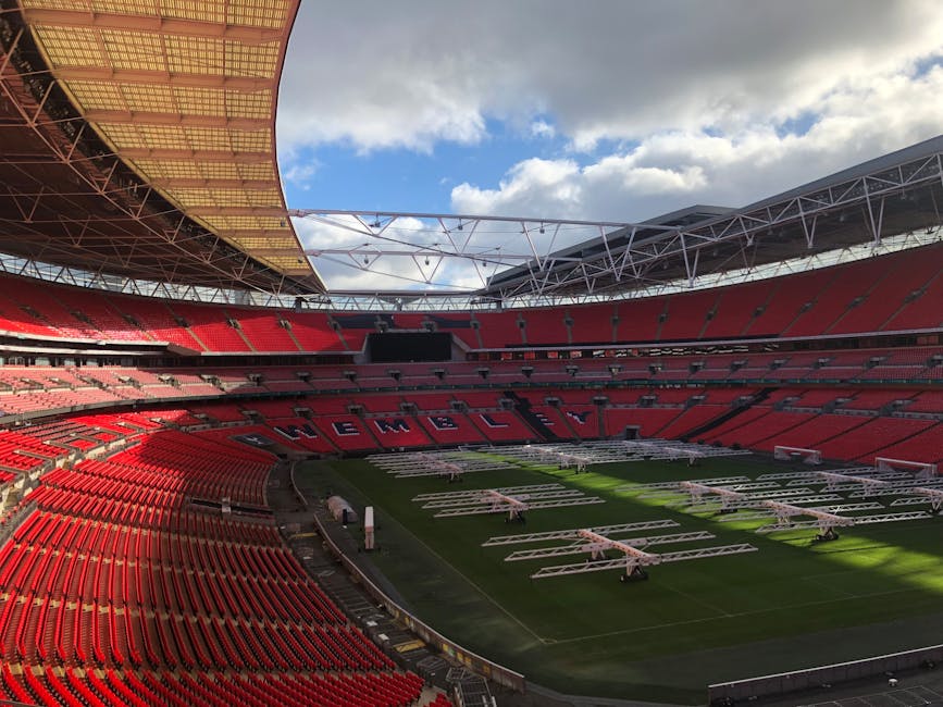 An expansive view of a large sports stadium with bright red seats arranged in multiple tiers surrounding the central grass pitch. The seats are mostly empty, and the word 'WELCOME' is spelled out across the middle section. The field is covered in white metal scaffolding structures, likely for a planned event, and some equipment and machinery are visible on the grass. The stadium roof features a modern, partially open design with a mix of metal trusses and a large, curved, gold-colored panel on the left side, allowing natural light to illuminate the interior. The background shows a partly cloudy sky with patches of blue. The scene suggests preparations for a large event such as a match or concert, demonstrating the capability for on-site staging and independent event handling outside standard venue operations, aligning with private rubbish removal or setup services for event day logistics. Rubbish Collection Twickenham may be involved in post-event clean-up or waste management for such large-scale gatherings.