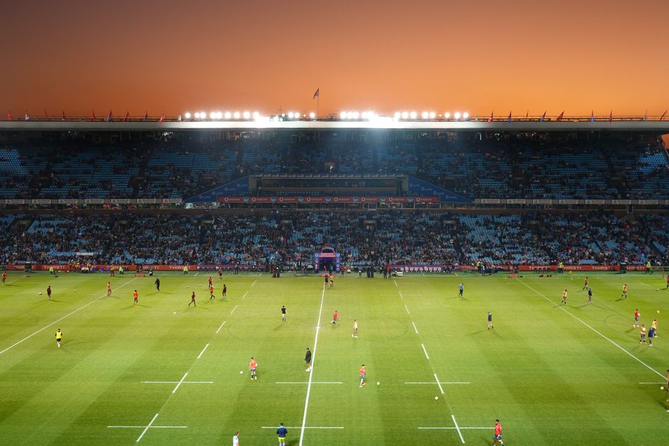 A wide view of a large, modern football stadium with a well-maintained green grass pitch marked with white lines. The stadium features blue seating sections on both the left and right sides, extending across multiple tiers. Above the seating areas, there are extensive roof structures made of metal and glass, supported by large steel trusses that create an open yet sheltered environment. The sky above is overcast, casting diffuse lighting over the scene, with no spectators or players visible on the field. Surrounding the pitch, the concrete walkways and support columns are visible, contributing to the overall structure. This scene represents an empty stadium prepared for a match, with a focus on the pristine condition of the grass and the architectural design of the roof, aligning with the context of large-scale sports venues where occasional maintenance or on-site clearance may be required, akin to private waste handling or refurbishment activities by professional rubbish removal services like Rubbish Collection Twickenham.